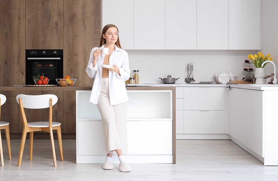 Una giovane donna vestita interamente di bianco beve un caffè in una cucina bicolore sui toni del bianco e del legno naturale, con pavimento in parquet bianco