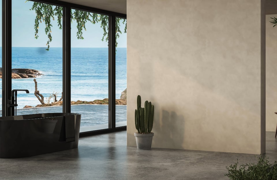 Ampia stanza da bagno affacciata, attraverso una parete vetrata, su una spiaggia. Le pareti interne sono in color écru e si vedono una vasca da bagno scura e un cactus in vaso