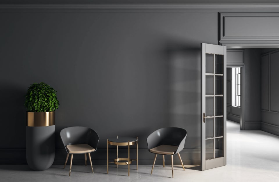 Lounge with grey armchairs and coffee table, next to a large grey vase with metal details, grey walls and a glass door of the same colour, leading into the rest of the house, where the walls are also grey