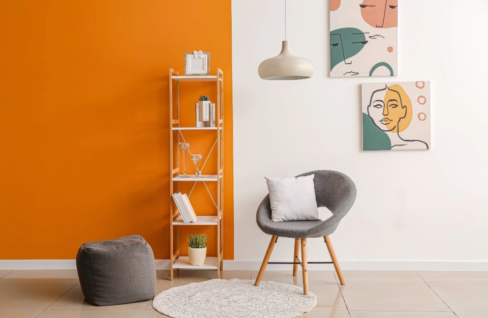 Stanza con parete bicolore (giallo ocra a sinistra, bianca a destra) con una piccola libreria a colonna in legno naturale con libri e oggetti, una poltrona circolare grigia in stile scandinavo, un pouf grigio, un lampadario a soffitto bianco in metallo bombato e due stampe alle pareti con illustrazioni sui toni del bianco, del giallo, del rosa e del turchese