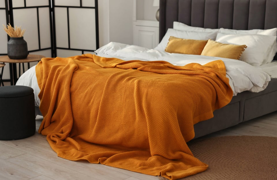 Letto matrimoniale con struttura grigio scura imbottita, lenzuola bianche, coperta color ocra, cuscini sui toni del giallo e, attorno, si intravedono un séparé in stile orientale, uno sgabello nero circolare, un tappeto rotondo e dei tavolini circolare in legno con strutture in metallo nero