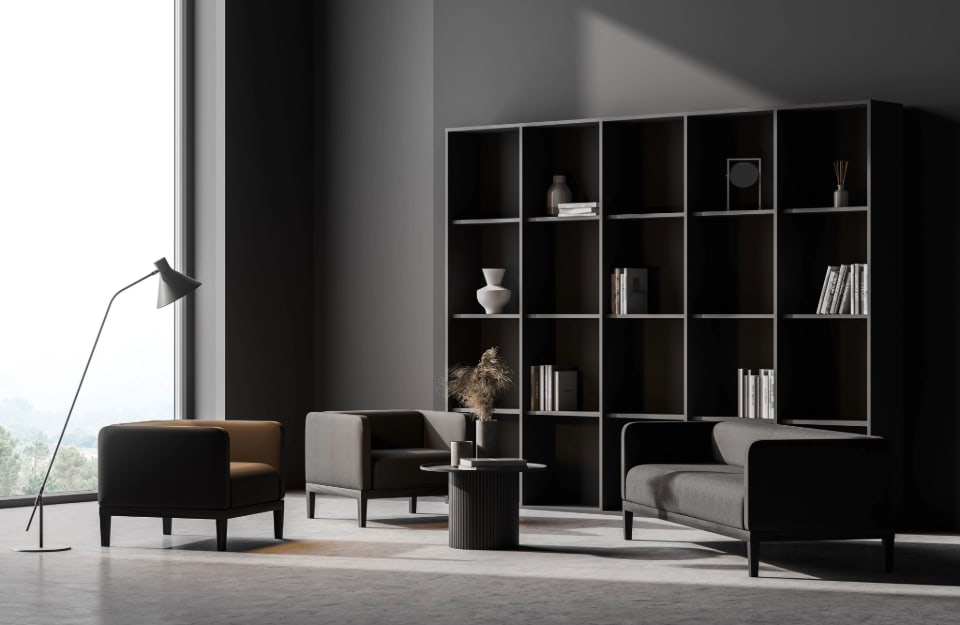 Angolo di un salotto in stile minimalista, tutto sui toni del grigio e del nero, con grande vetrata, poltrone e sofa dalle linee essenziali, grande libreria nera con pochi libri e oggetti, tavolino da caffè scuro con base cilindrica e piano circolare, lampada da terra