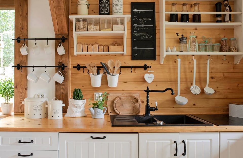 Area di una cucina in stile rustico, con parete in legno, mobili in legno naturale e bianco, molte mensole con spezie, vasetti, tazze e attrezzi da cucina, maniglie e rubinetto in metallo nero, lavagnetta appesa alla parete e piantine sul pianale