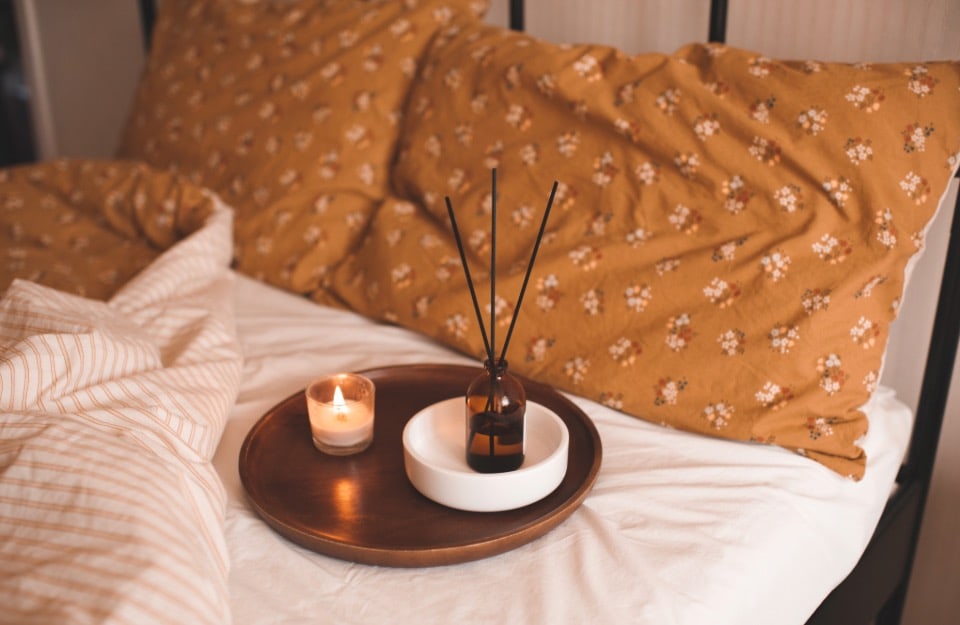 Detail of an unmade bed in tones of light brown, with a tray containing a scented candle and an ampoule essence diffuser