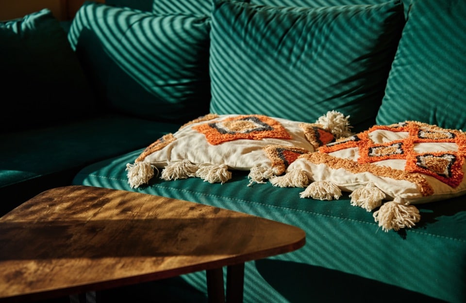 Detail of a petrol green sofa with ethnic cushions next to a coffee table of with an irregular dark natural wood top. A play of shadows can be seen on the sofa