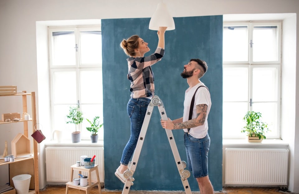 Una joven pareja está decorando su casa: ella está subida a una escalera y atornilla la bombilla de una lámpara de araña mientras él sujeta la escalera. La habitación es luminosa y entre dos ventanas hay una pared pintada de azul ballena