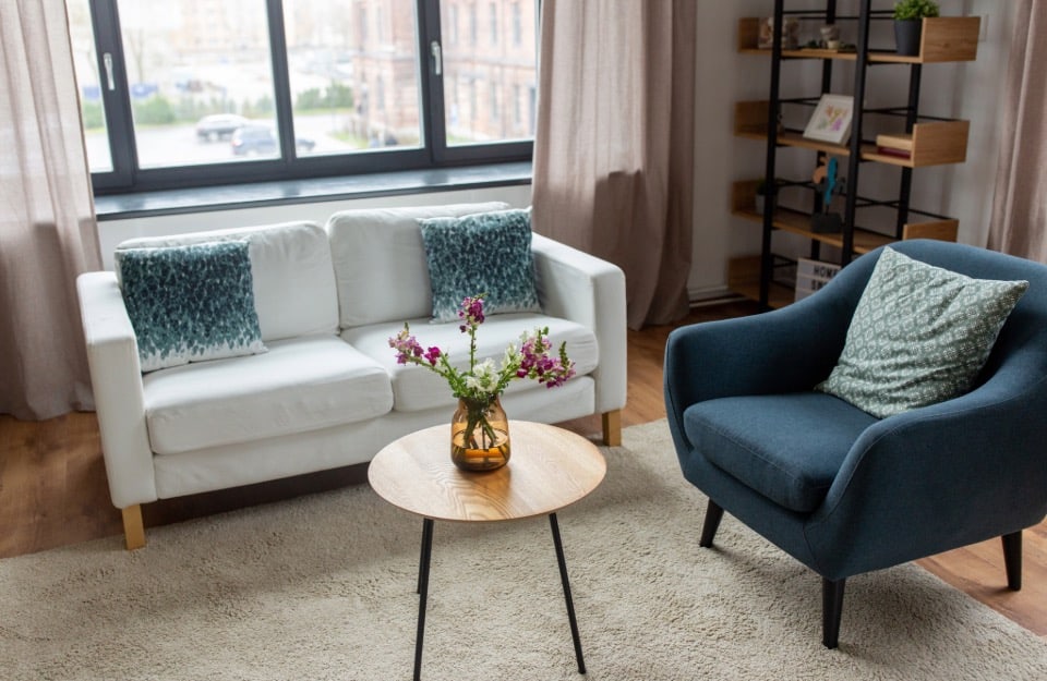 Rincón de un salón con sofá blanco, sillón azul, mesa de centro con un jarrón de flores encima, alfombra y, al fondo, una ventana y una librería.