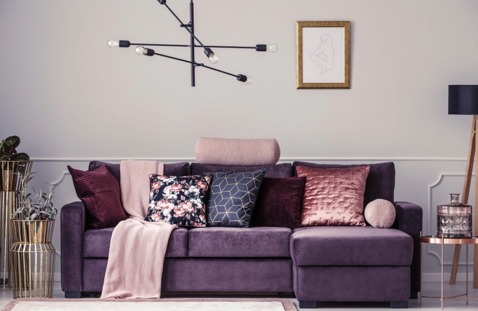 Soggiorno elegante e moderno. L'elemento principale è un divano sezionale viola adornato con vari cuscini decorativi di diverse texture e colori, tra cui motivi floreali, geometrici e solidi. C'è una coperta rosa chiaro drappeggiata sul bracciolo del divano. Sopra il divano, c'è una moderna lampada nera con bulbi esposti e un disegno minimalista incorniciato di una figura seduta. A sinistra del divano, c'è un supporto per piante dorato con piante in vaso, e a destra, c'è un tavolino laterale con un vaso di vetro e una lampada nera con paralume su un treppiede di legno