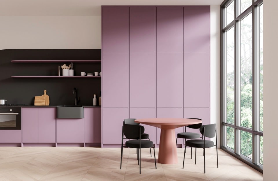 Cucina moderna con zona pranzo. La cucina presenta armadietti di colore lilla chiaro con una finitura opaca, inclusi armadietti alti, armadietti bassi e una sezione con scaffali aperti. La zona del backsplash e il piano di lavoro sono neri, e c'è un lavello nero. La zona pranzo include un tavolo rotondo con base e piano di colore rosa, circondato da tre sedie nere. La stanza è ben illuminata grazie a grandi finestre che vanno dal pavimento al soffitto sul lato destro. Il pavimento è in legno chiaro disposto a spina di pesce