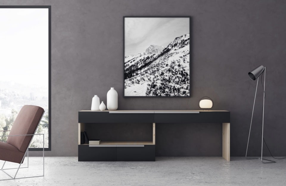 Tavolo console nero contro una parete di color grigio lavagna con nuance violacee, con una grande fotografia in bianco e nero di un paesaggio montano innevato appesa sopra. Sul tavolo console ci sono tre vasi decorativi bianchi di diverse dimensioni sul lato sinistro e una piccola lampada rotonda sul lato destro. A sinistra del tavolo console, c'è una grande finestra con vista su un paesaggio nebbioso e alberato all'esterno. Davanti alla finestra, c'è una sedia imbottita marrone con una struttura metallica. A destra del tavolo console, c'è una lampada da terra alta e nera con uno stelo sottile e angolato