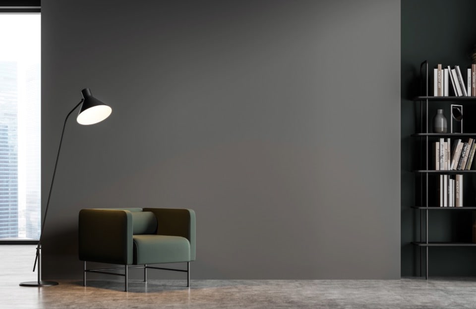 Spazio interno moderno e minimalista. L'elemento principale è una poltrona verde scuro con un design semplice e squadrato. Accanto c'è una lampada da terra alta e nera con un paralume interno bianco. Sul lato destro della scena si trova una libreria in metallo nero con vari libri e alcuni oggetti decorativi. Lo sfondo è una parete grigio tartufo e c'è una grande finestra a sinistra che permette alla luce naturale di entrare nella stanza