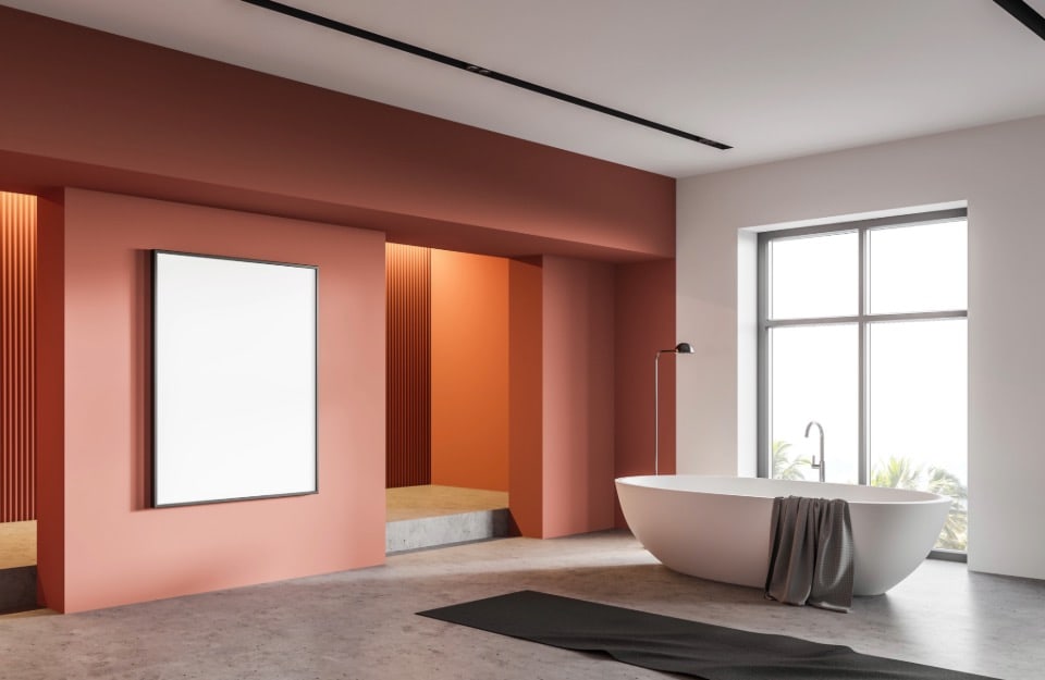 Un bagno moderno dal design minimalista. La stanza presenta una vasca bianca indipendente con un asciugamano grigio appoggiato sul bordo, situata vicino a una grande finestra che fa entrare molta luce naturale. Le pareti sono dipinte in una combinazione di colori terracotta calda e bianco