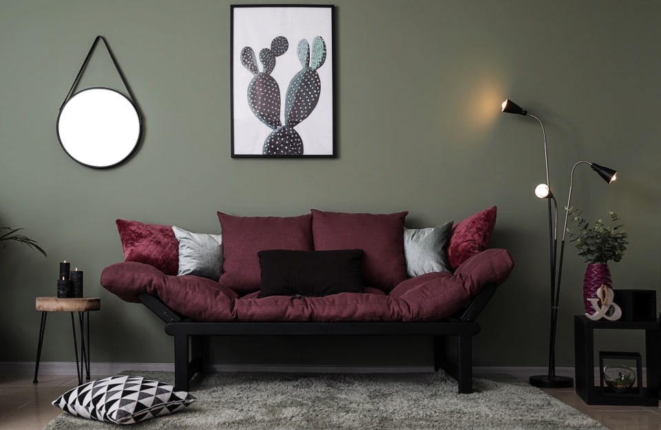 Soggiorno moderno con una parete verde scura, divano futon color borgogna con cuscini neri, grigi e borgogna. Uno specchio rotondo con una cornice e una cinghia nere è appeso alla parete sulla sinistra.
Un quadro incorniciato di un cactus con uno sfondo bianco appeso sopra il futon.
Un piccolo tavolino laterale di legno con gambe in metallo nero sulla sinistra del futon.
Una lampada da terra con tre luci regolabili sulla destra del futon