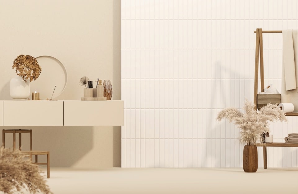 Bagno minimalista. Sul lato sinistro c'è una mensola sospesa con vari oggetti decorativi tra cui un vaso con fiori secchi, una candela, uno specchio rotondo e alcuni piccoli contenitori. Sul lato destro, c'è una mensola a scala in legno che sostiene un asciugamano arrotolato, un asciugamano piegato e un piccolo cestino. C'è anche un vaso alto con erba delle pampas secche sul pavimento accanto alla mensola. Lo sfondo presenta una parete con piastrelle bianche verticali