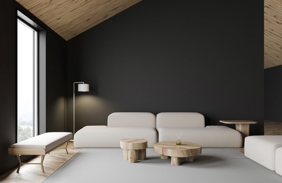 Soggiorno moderno e minimalista con un alto soffitto inclinato in legno e pareti scure. La stanza presenta una grande finestra sul lato sinistro, che permette alla luce naturale di entrare. Vicino alla finestra c'è una panca imbottita bianca, mentre al centro della stanza si trova un divano bianco a basso profilo con cuscini. Davanti al divano ci sono due piccoli tavolini rotondi in legno. Una lampada da terra con un paralume bianco è posizionata accanto al divano, e un piccolo tavolino laterale in legno è collocato alla fine del divano. Il pavimento è una combinazione di legno chiaro e un tappeto chiaro