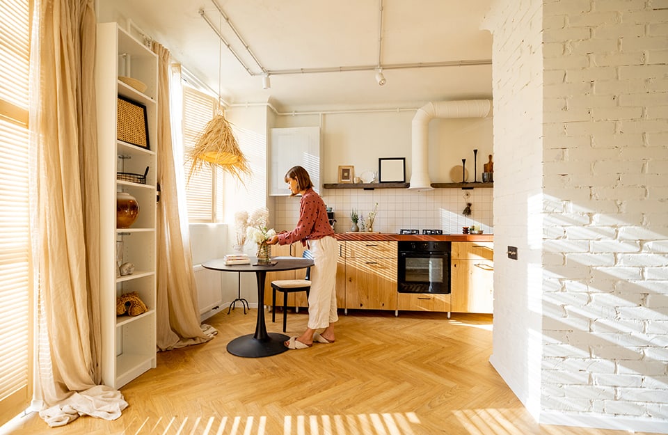 Una luminosa e accogliente cucina e sala da pranzo. La cucina presenta mobili in legno, un forno nero e un backsplash di piastrelle bianche. C'è un piccolo tavolo da pranzo rotondo con una base nera, e una donna sta sistemando dei fiori su di esso. La stanza ha un pavimento in legno chiaro con motivo a spina di pesce. Alla sinistra, c'è una alta libreria bianca con vari oggetti decorativi, tra cui cesti, vasi e libri. Lo spazio è ben illuminato dalla luce naturale che entra attraverso le grandi finestre con tende beige. Sopra il tavolo da pranzo, c'è un lampadario unico fatto di fibre naturali. Le pareti sono dipinte di bianco, e c'è una parete in mattoni bianchi sul lato destro dell'immagine